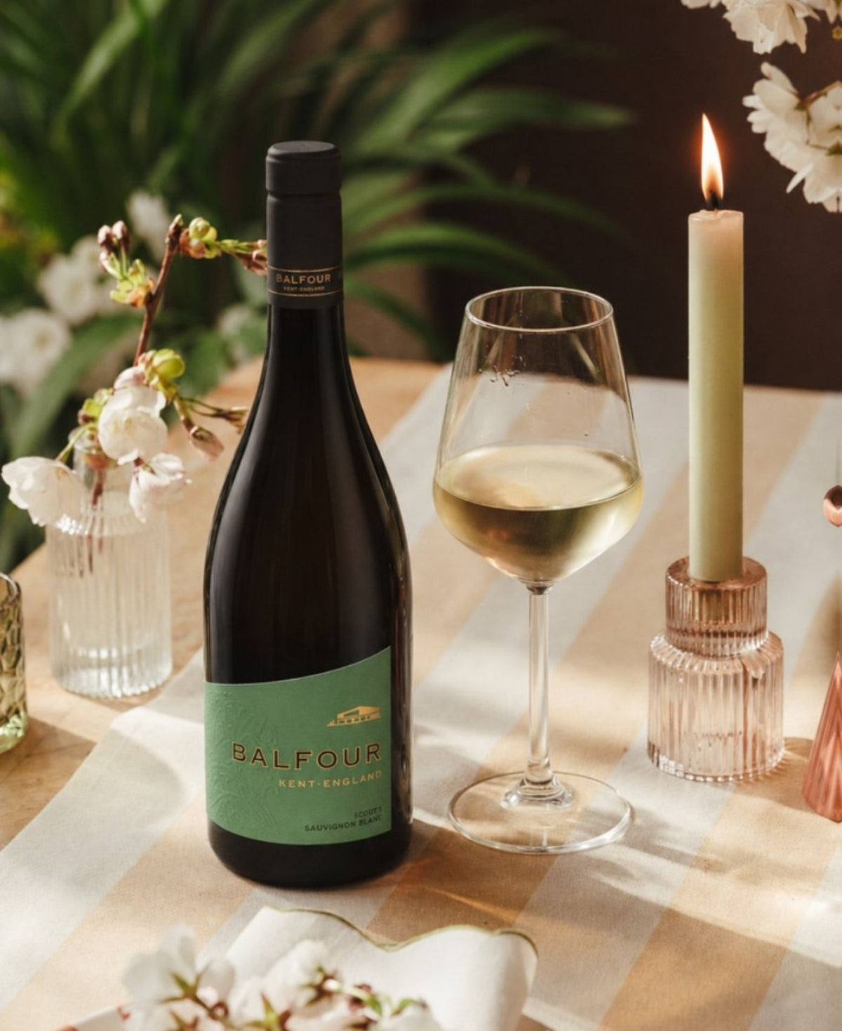 A bottle of Balfour white wine stands on a table beside a partially filled wine glass, a lit candle in a glass holder, and small floral arrangements in a softly styled indoor setting.