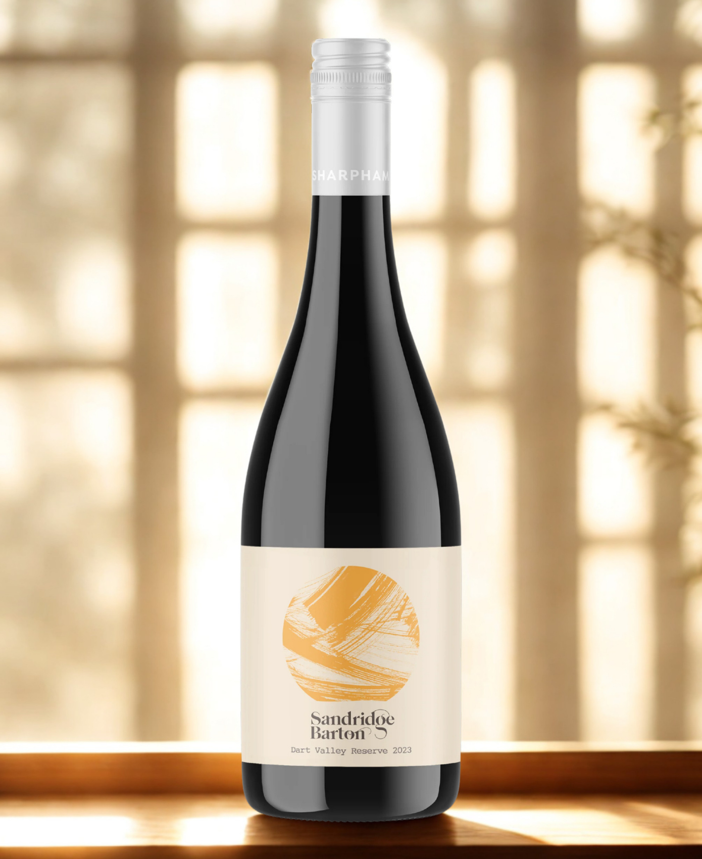 A bottle of Sandridge Barton wine with a cream label featuring a gold abstract design, set against a warm, softly blurred window background.