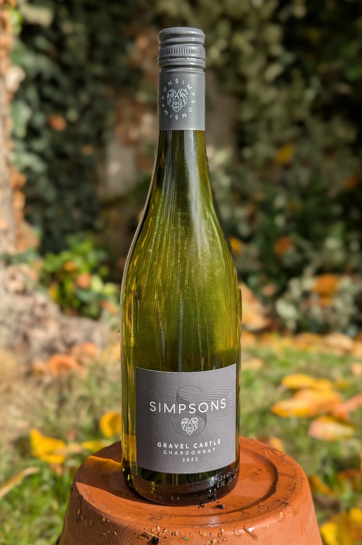 A bottle of Simpson's Gravel Castle white wine, in a green glass bottle with a gray label and silver screw cap, displayed on a terracotta pot against a blurred garden background.