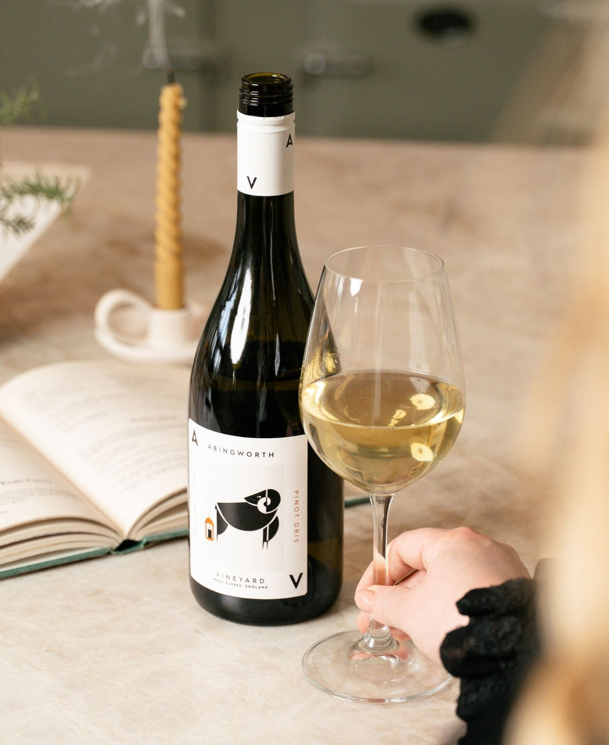 A bottle of Abingworth Pinot Gris wine next to a glass of pale golden wine, with a hand holding the glass on a beige tabletop.