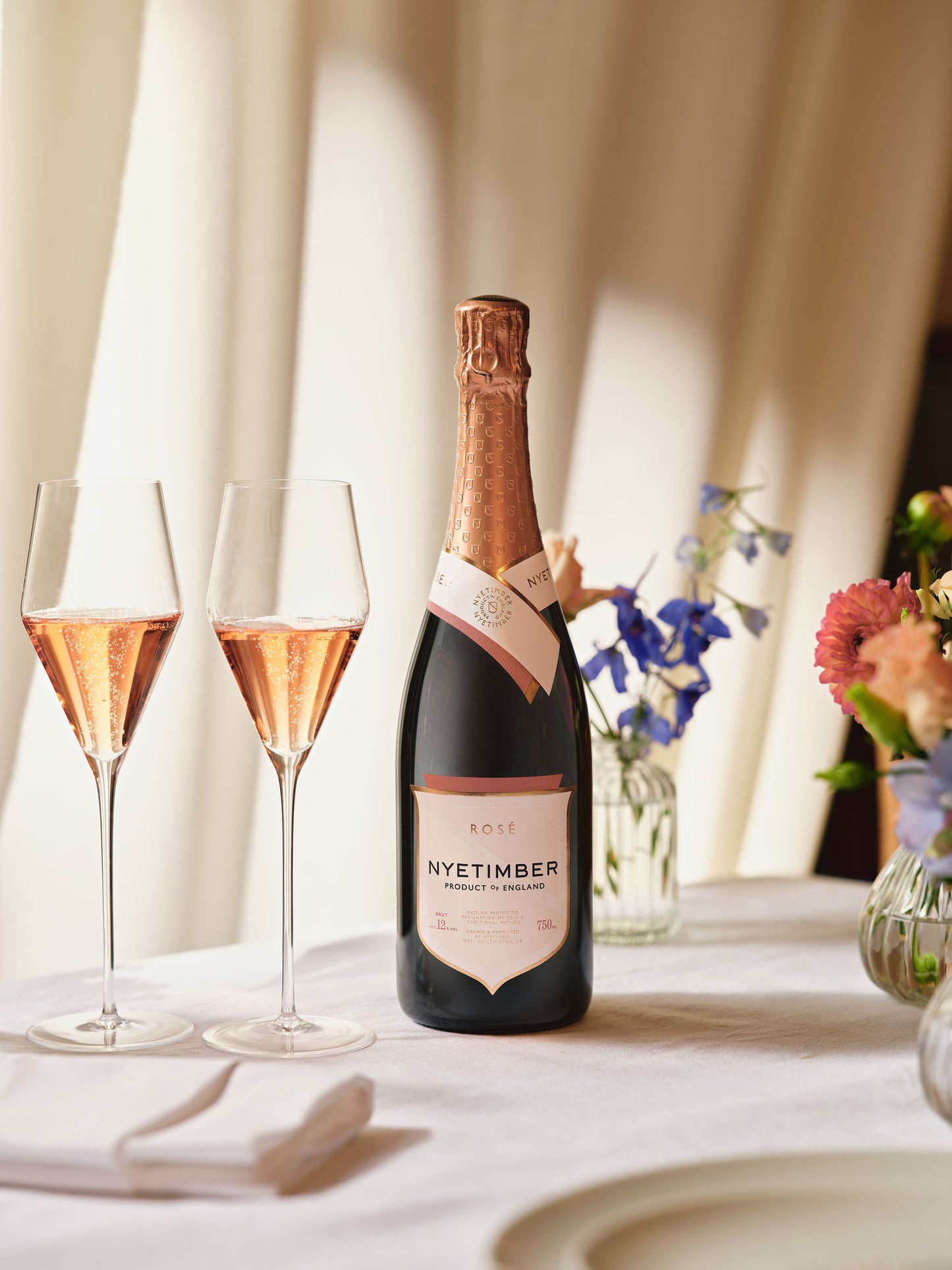 Nyetimber rose champagne bottle with two glasses on a table with flowers in the background